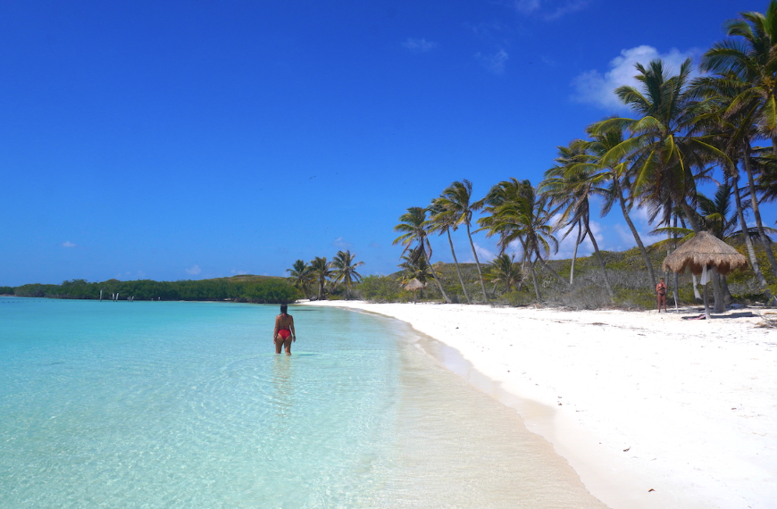 Mujer en traje de baño rojo caminando por la orilla de una playa de arena blanca, aguas cristalinas turquesas y palmeras bajo un cielo azul despejado.