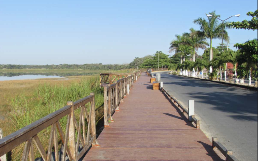 Malecón de madera frente a una laguna costera con palmeras y carretera en un pueblo pesquero de Yucatán, México.
