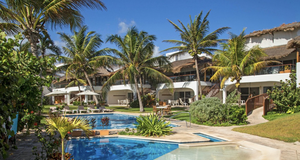 Complejo residencial de lujo con alberca, jardines tropicales y villas con techos de palapa frente al mar.