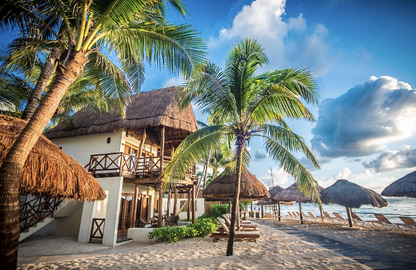 Luxury beachfront hotel with thatched palapa roofs in the Caribbean, surrounded by palm trees and beach loungers.