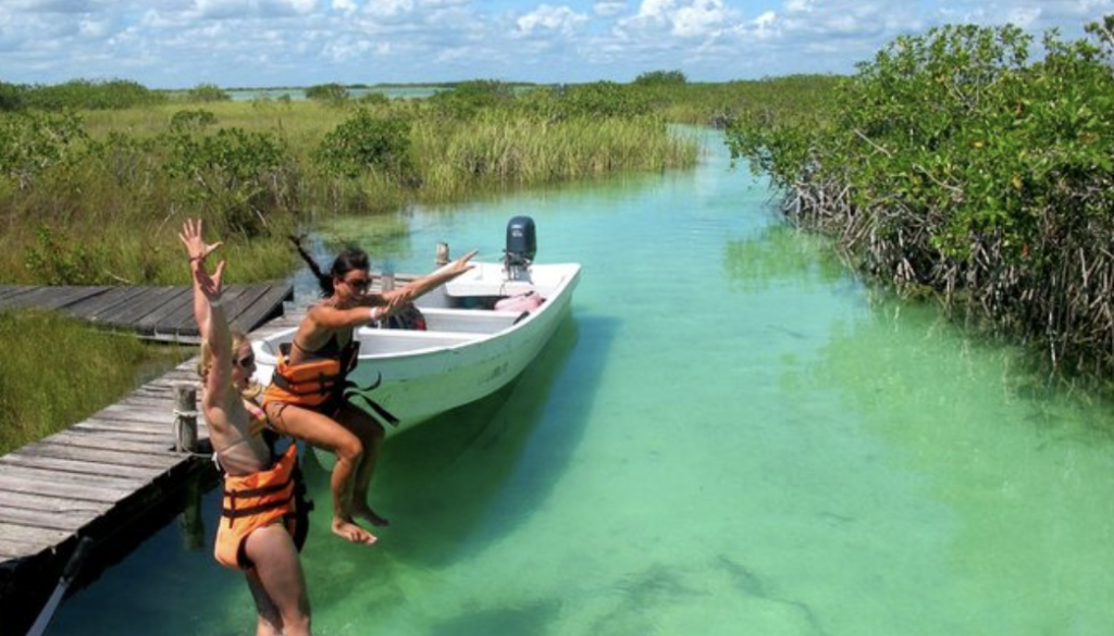 Tourists jumping into crystal clear water from a wooden dock in Sian Ka'an canals, Muyil, with a small boat nearby.